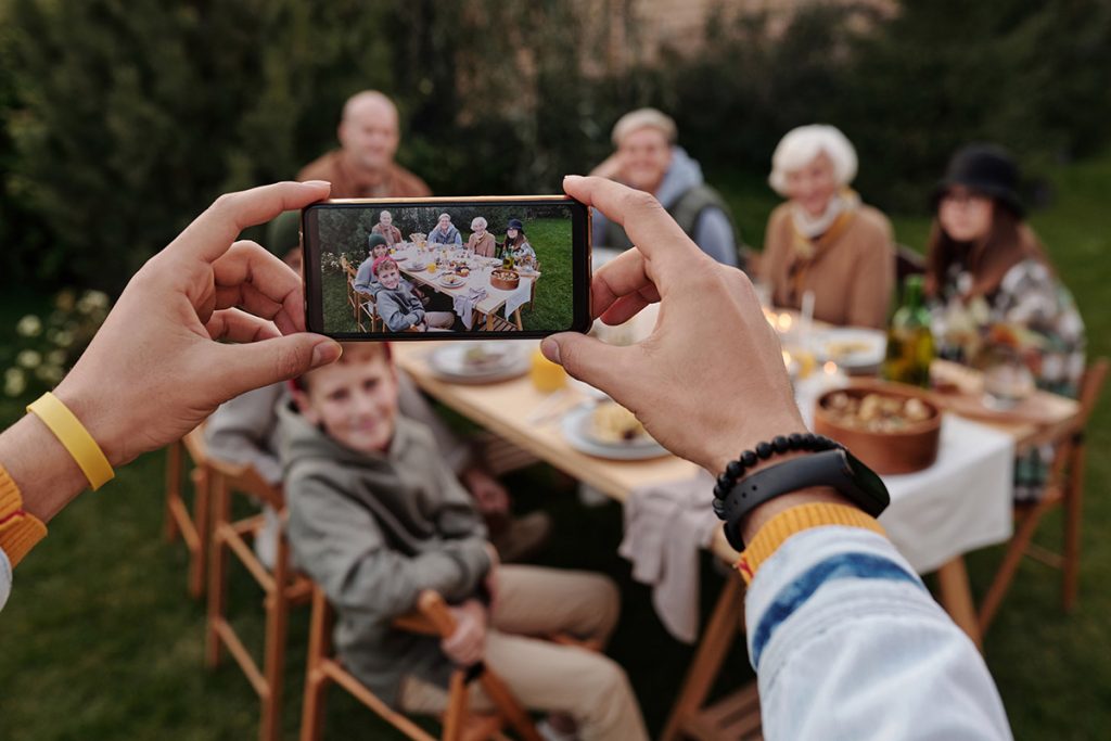 Someone taking a photo on their phone of a family sitting at a table outside.